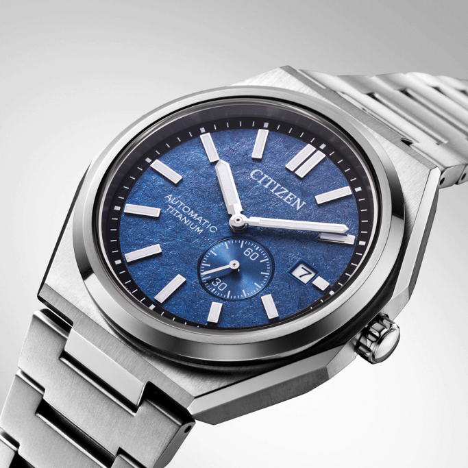 A close-up studio shot of a Citizen automatic watch. It has a blue textured face, silver markers, and a stainless steel band. The focus is on the watch face and its design.
